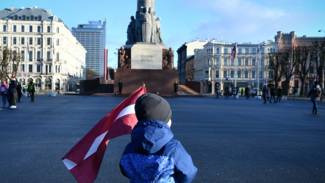 bērns nes Latvijas karogu, fonā Brīvības piemineklis