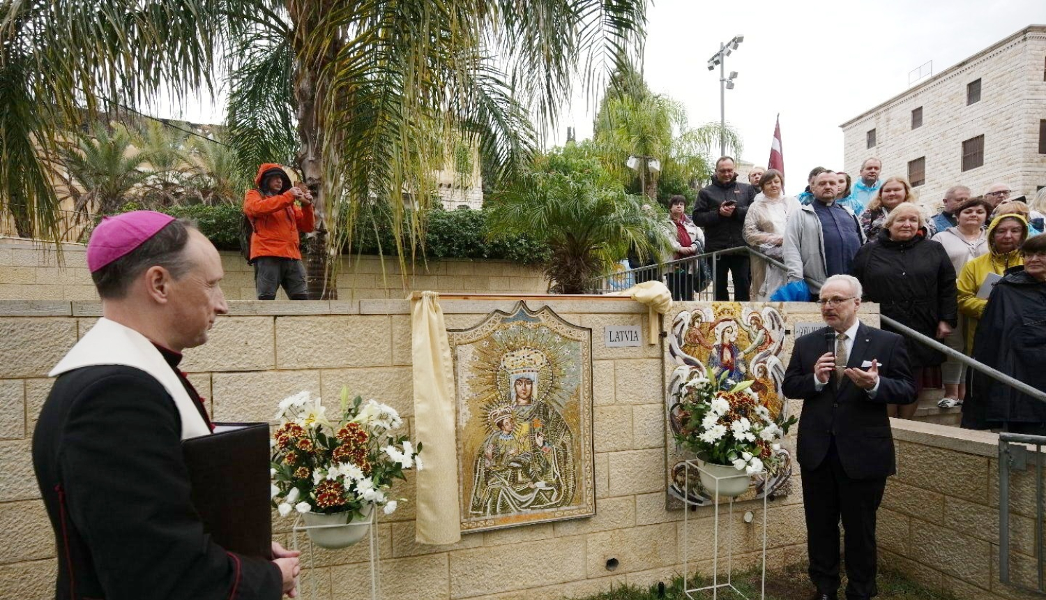 Garīdznieks un Valsts prezidents stāv pie mozaīkas, kas izvietota uz mūra sienas
