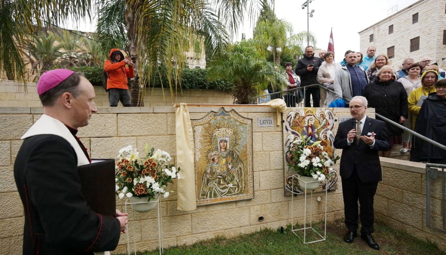 Garīdznieks un Valsts prezidents stāv pie mozaīkas, kas izvietota uz mūra sienas